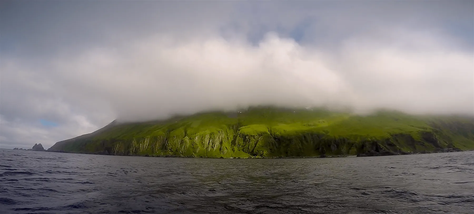 Lush green mountains are covered in clouds. Dark ocean water is seen. 