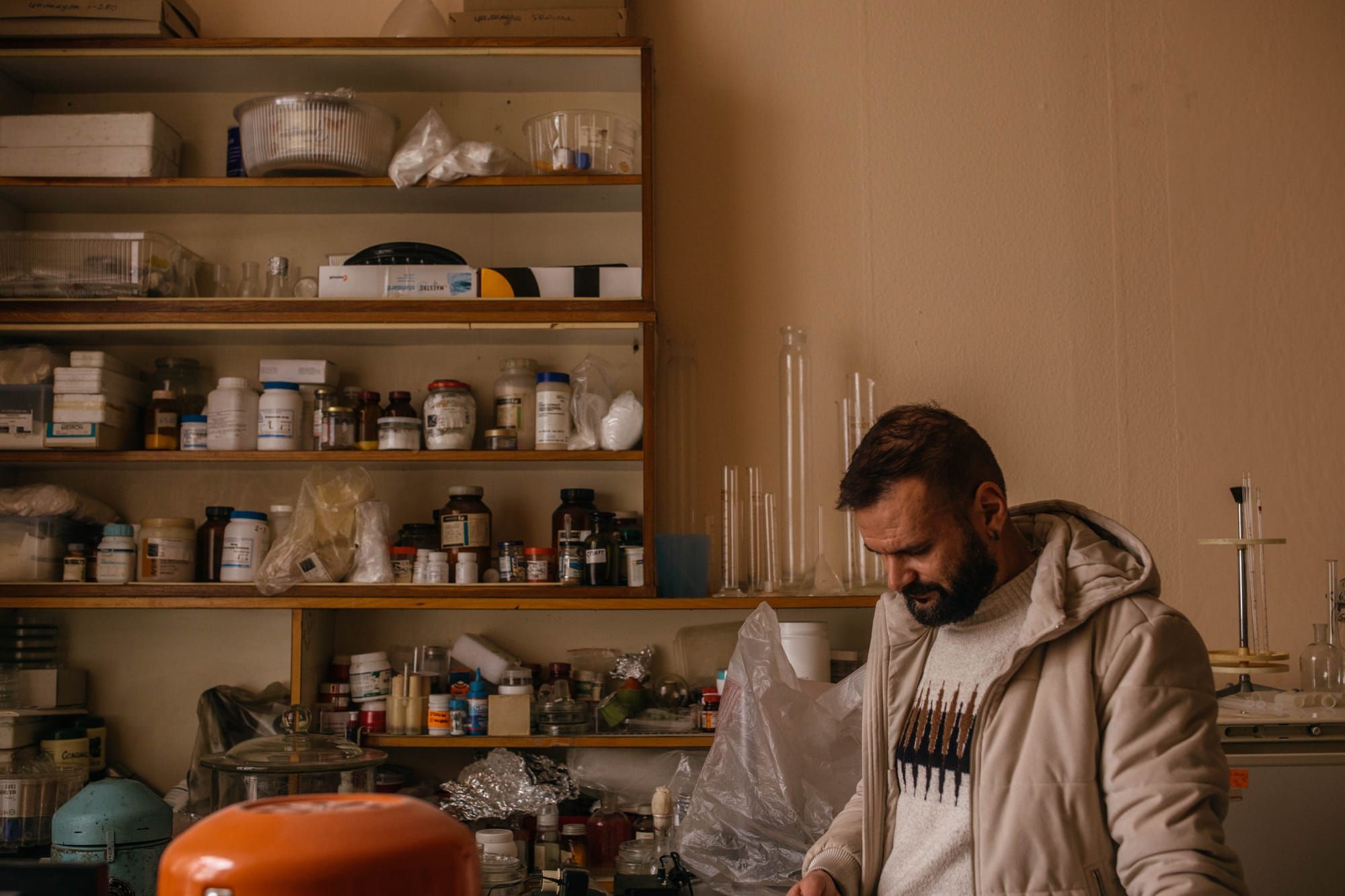A brown man in a jacket and sweatshirt conducts research in a laboratory. Behind him are shelves stocked with bottles.