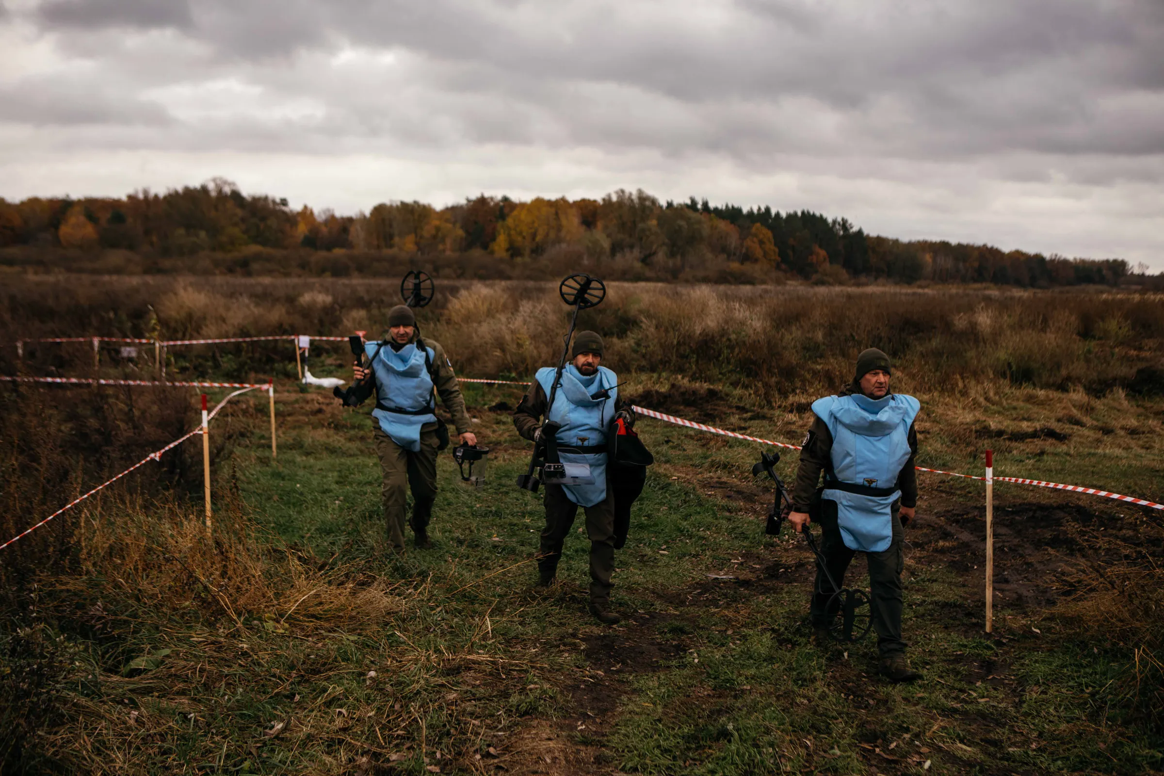 Three men in black clothing and blue protective gear walk along a barricaded path beside an agricultural field, carrying detection devices.