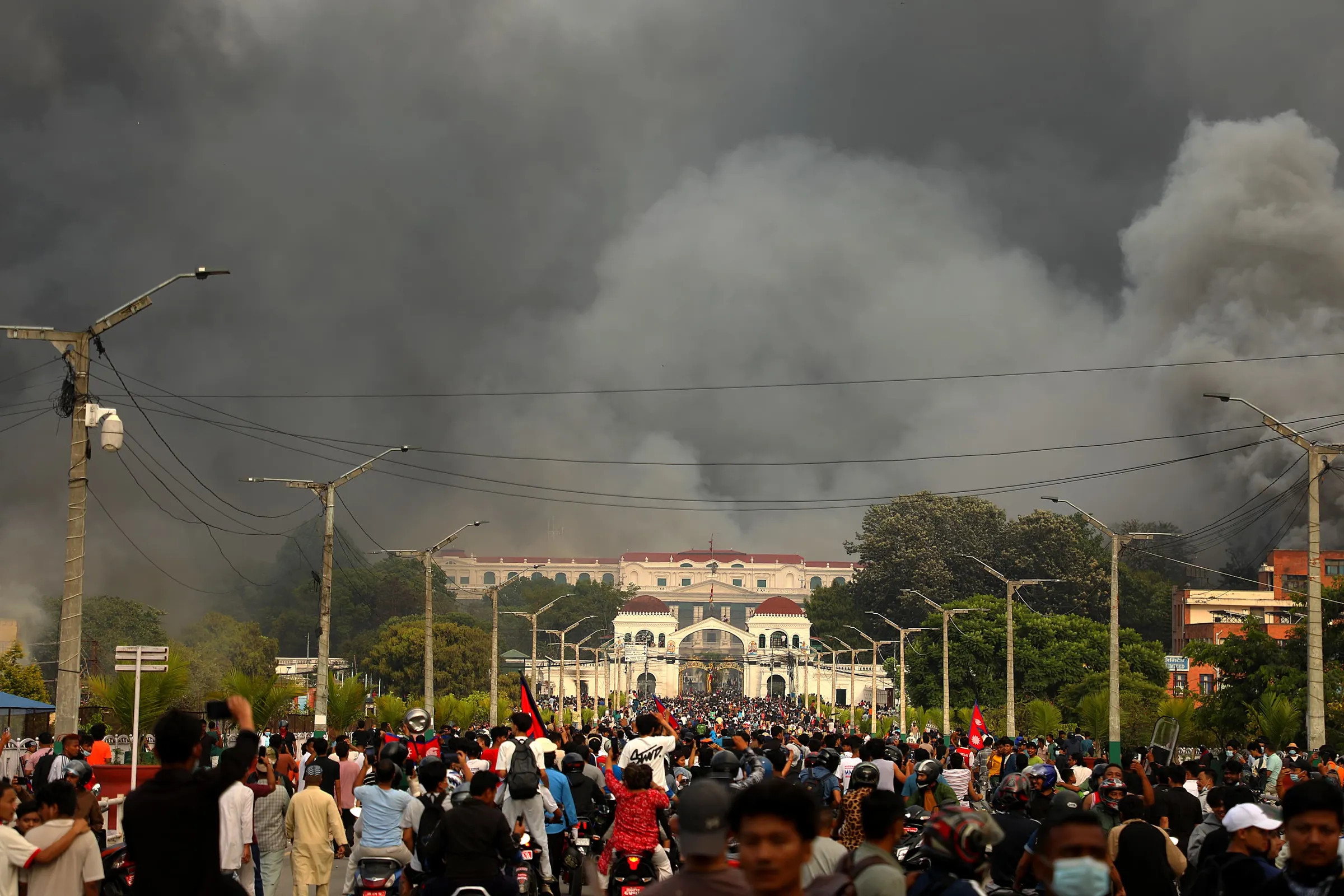 People holding red and black flags stand outside a large government building, with some taking photos as smoke rises and spreads across the sky above it.