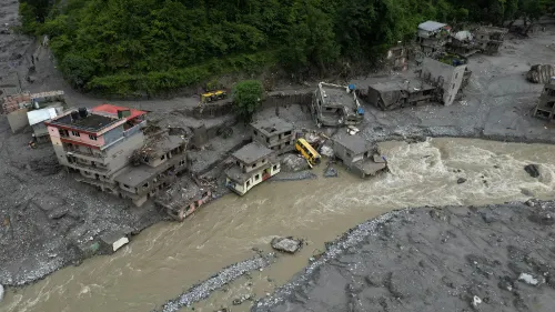 A gush of floodwater flows through the area, with homes buried under mud. A few houses remain on the other side, but most are surrounded by debris.