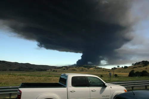 A vehicle drives down a highway with a dark cloud of black smoke engulfing the sky in the background.