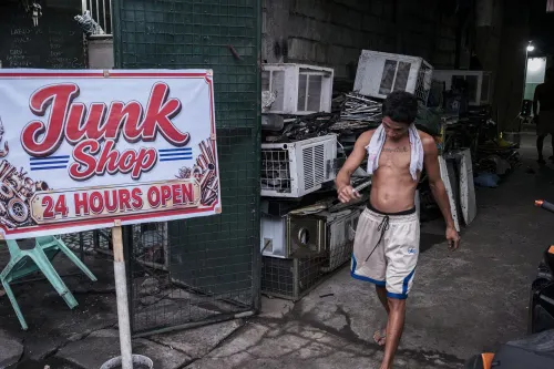A shirtless man with a towel draping over his shoulders, wearing white board shorts and flip-flops, walks past dismantled air conditioners and a sign that writes, "Junk Shop 24 HOURS OPEN".