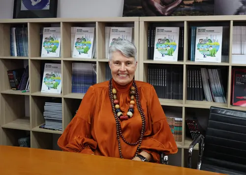 Debra Roberts sits at a wooden desk in front of bookshelves filled with books. She has short gray hair and wears an orange silk blouse and a large bead necklace. 