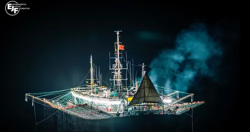 A brightly lit squid fishing vessel at night, it's deck lined with nets and lights stretching into the dark sea.