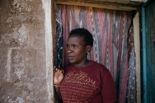 Lucy Kagure stands in the doorway of ther home in Nanyuki, Kenya, wearing a maroon T-shirt, her face marked by a weary expression.