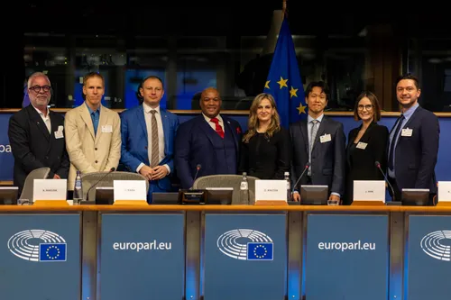 The members pose with other attendees during a conference on nanoplastics at the European Parliament, smiling at the camera, in front of a blue European Union flag.