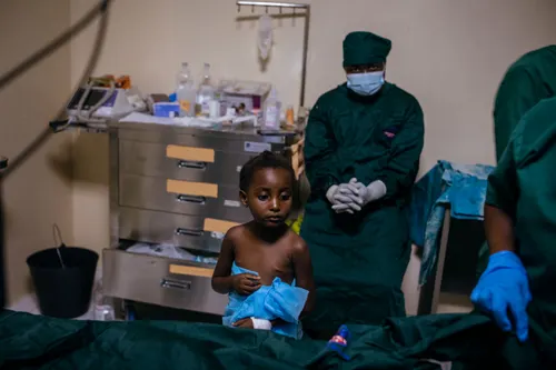 A four-year-old girl wrapped in a blue hospital sheet stands beside a hospital bed. She appears apprehensive.