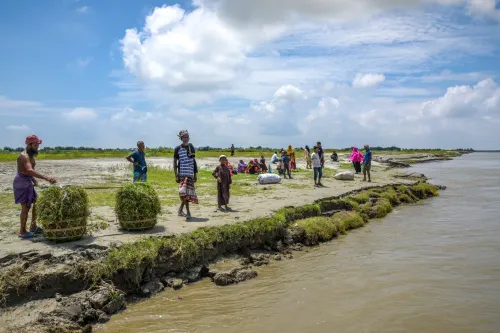 Men, women, and children wait on the shore of a water body, with two large baskets filled with fodder nearby.