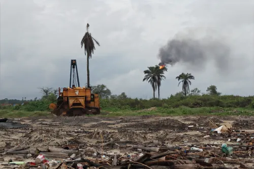 Behind a dirty beach is heavy machiney and a gas flare among a forest.