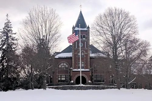 A snow-covered University of New Hampshire building stands with an American flag flying in front, surrounded by dried pine trees