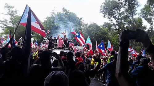 A crowd holding flags participating in a demonstration with flags and plumes of smoke wafting upwards.  