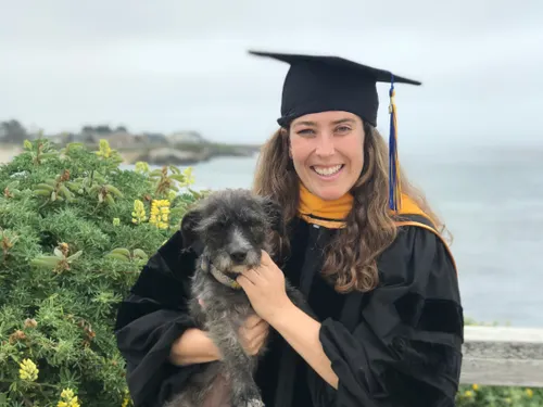 Kate wears her graduation cap and gown while holding a dog in her arms. Both pose for the camera.