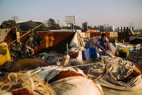 A fisherman in a plaid shirt sits among his tangle of nets at a pier.