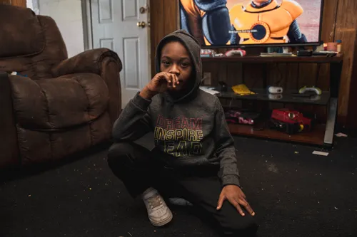 A Black boy with a Black hoodie with the words "DREAM INSPIRE LEAD" sits in front of a television and a brown couch.