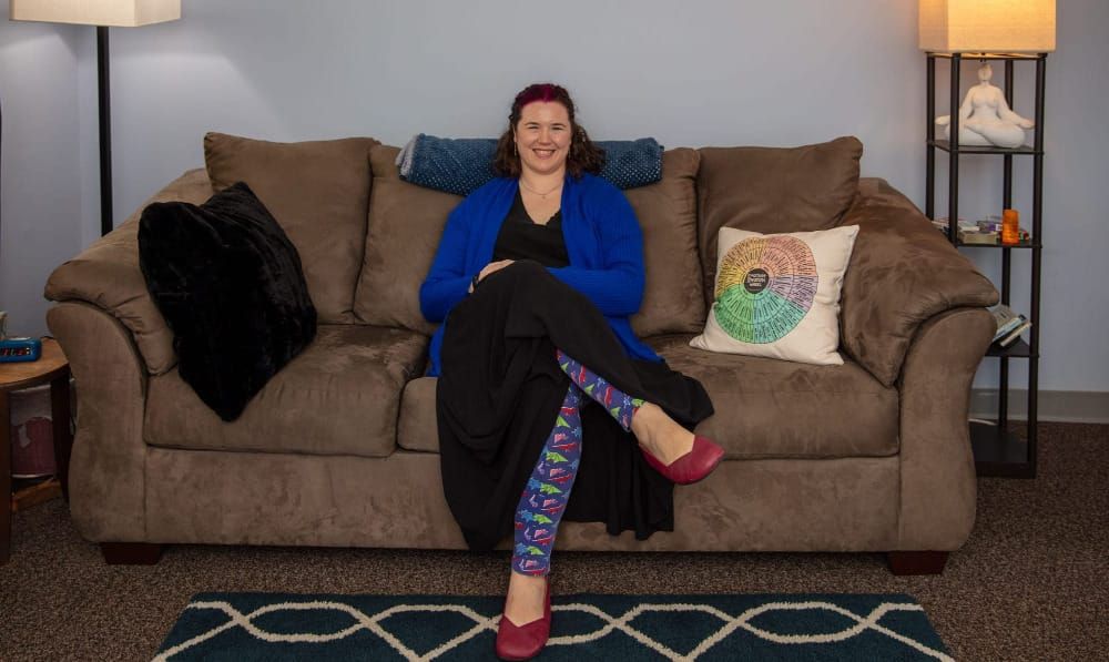 A light-skinned woman sits on a brown couch with her legs crossed at the knees.