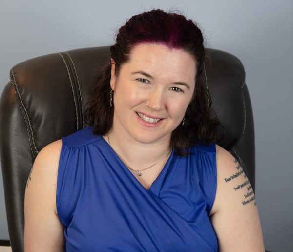 A light-skinned woman sits smiling at the viewer. She is wearing a blue sleeveless shirt. 
