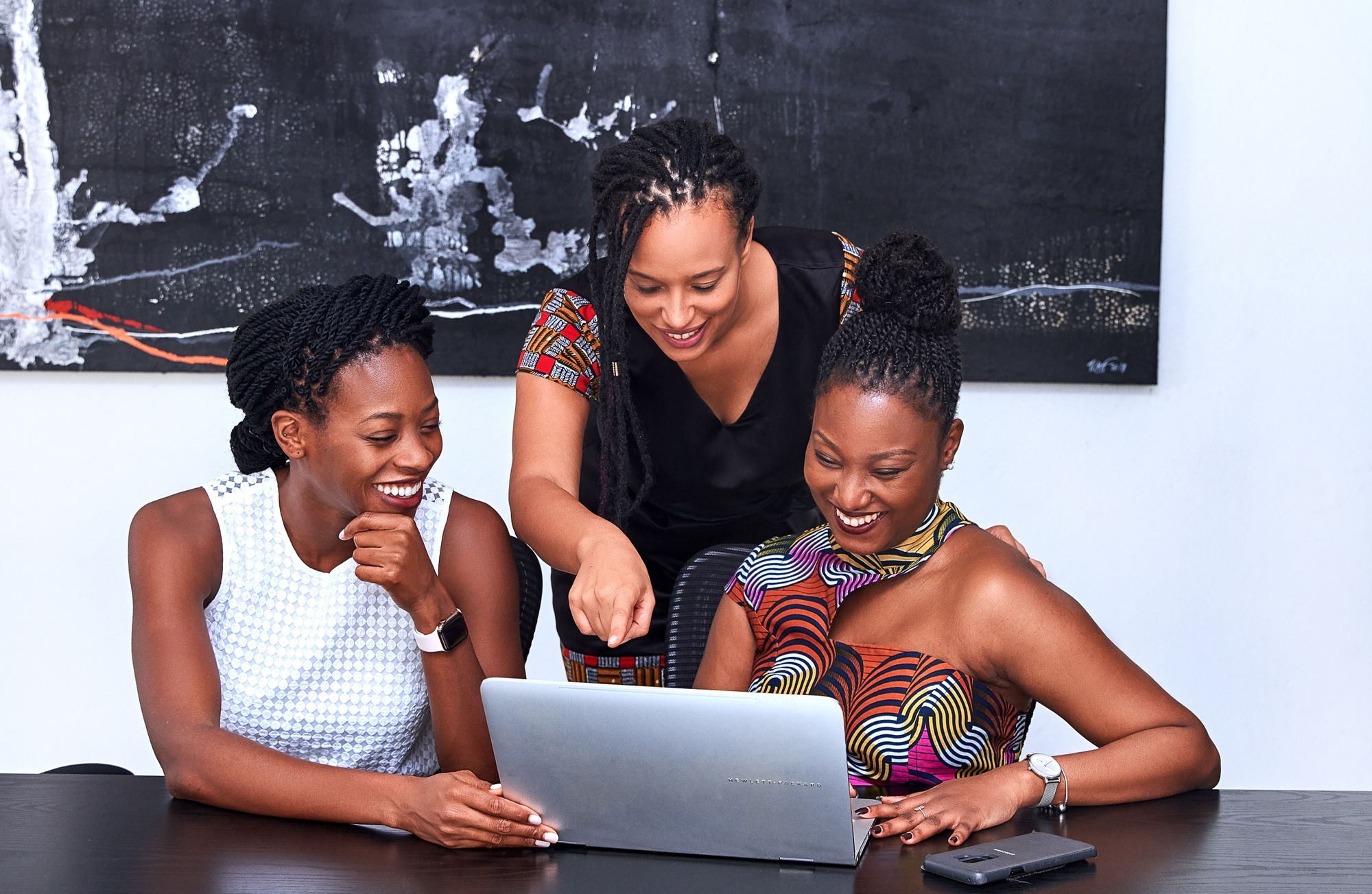 Três mulheres sorriem enquanto olham para notebook apoiado em mesa escura para ilustrar escolha por site de vaquinha. A primeira está sentada à esquerda e é negra de pele escura, com cabelo trançado preto para trás, usa vestido branco e olha para o notebook branco à sua frente. A segunda está em pé, é negra de pele clara, usa cabelo trançado solto, usa vestido preto com mangas vermelhas e aponta para a tela do mesmo notebook enquanto sorri. A terceira está sentada à esquerda, é negra de pele escura, com cabelo estilo coque, usa vestido de estampa colorida e sorri enquanto digita no mesmo notebook. O fundo é neutro, com parede branca e quadro preto.