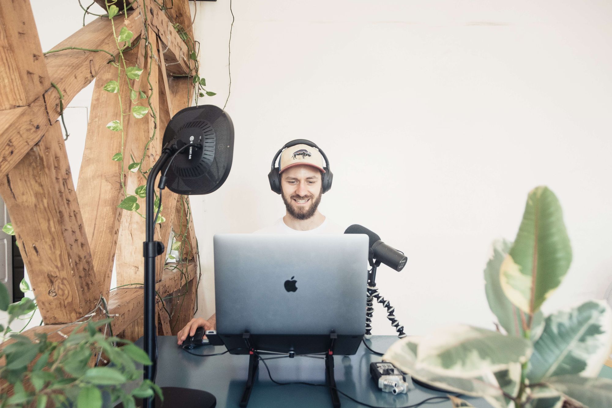 homem branco e de barba em uma mesa de gravação de podcat, ao lado esquerdo há uma estrutura vazada, de madeira e diversas plantas ao redor