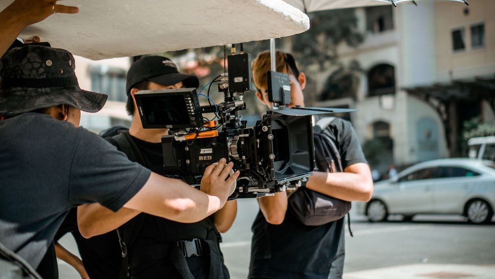 grupo de três homens fazendo filmagem em uma rua, eles seguram uma câmera profissional
