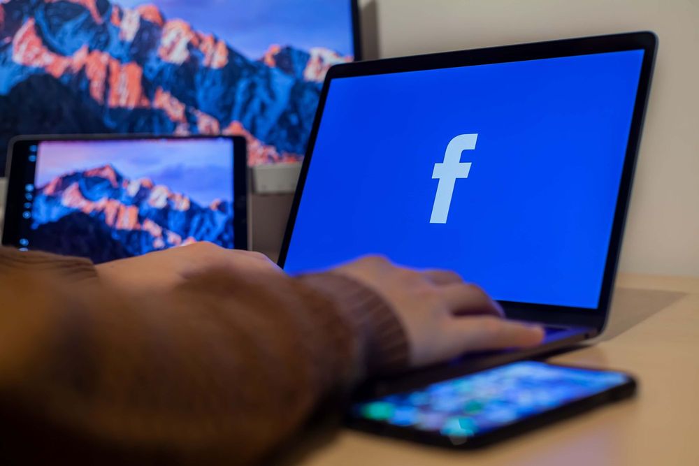 detalhe de um braço sobre um notebook, na tela, toda em azul, o logo do facebook, a letra f, na cor branca
