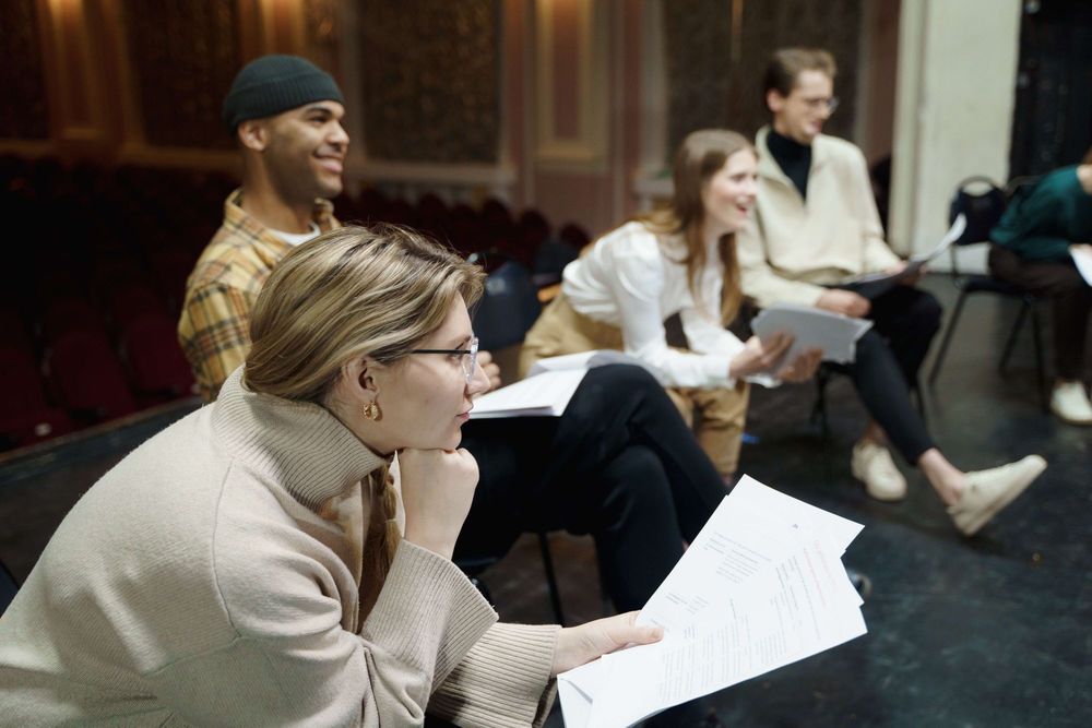 Grupo de pessoas sentadas em cadeiras sobre um palco de teatro. Elas seguram folhas na mão.