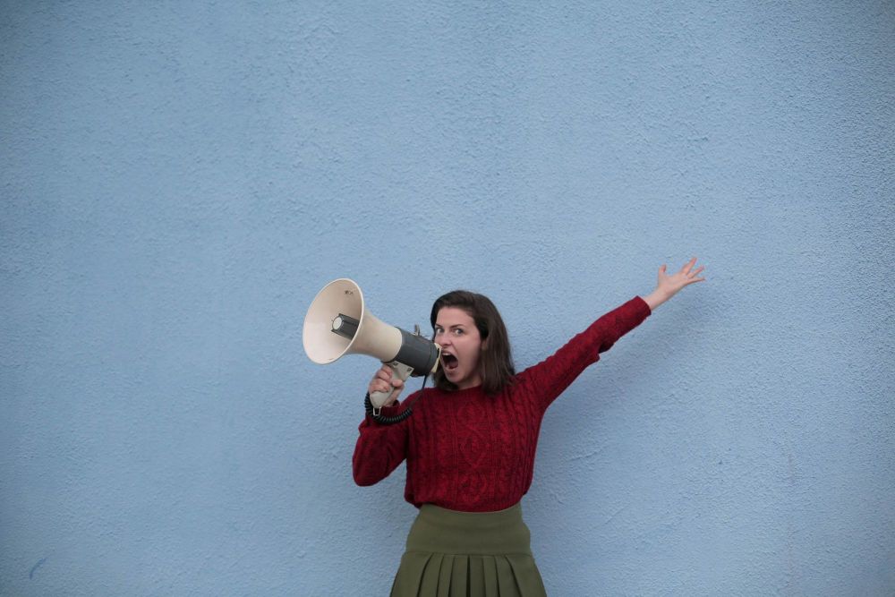 Mulher branca de saia verde e camisa vermelha, de mangas longas. Ela segura uma megafone próximo à boca. 