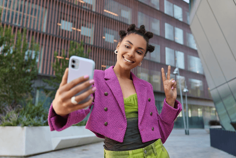 Mulher negra com casaco rosa e roupa verde, pousa para frente de um iPhone com capa lilás fazendo o sinal de paz e amor