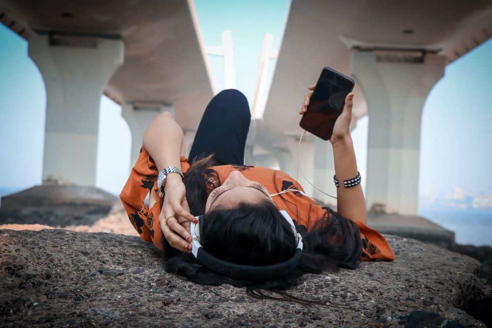 Foto em dia claro de uma mulher sobre uma pedra, embaixo de de uma estrutura em concreto de uma ponte. Ela usa fones d  