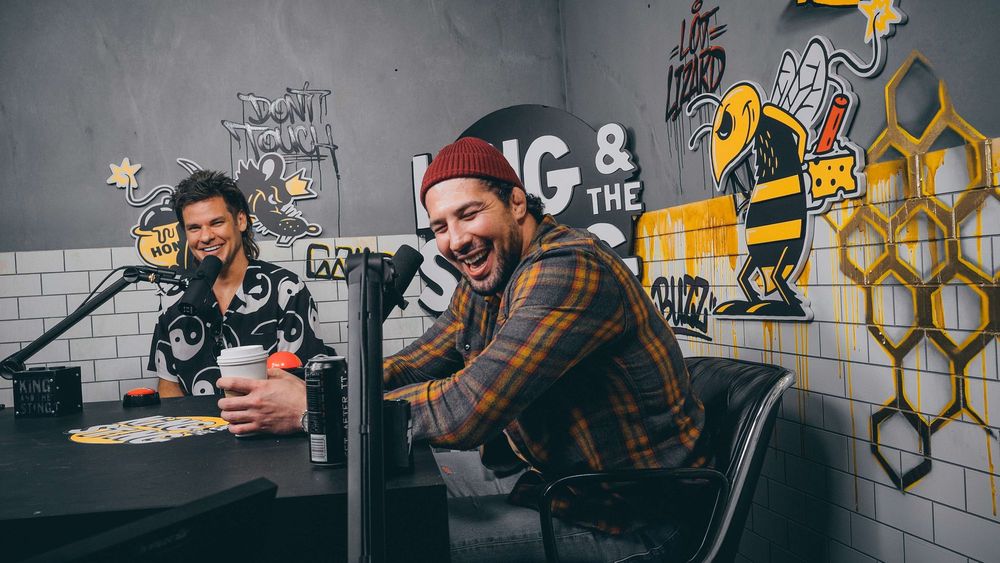 Foto de dois homens sentados junto a uma mesa com microfones. Eles usam roupas estampadas e o cenário ao fundo é colorido.