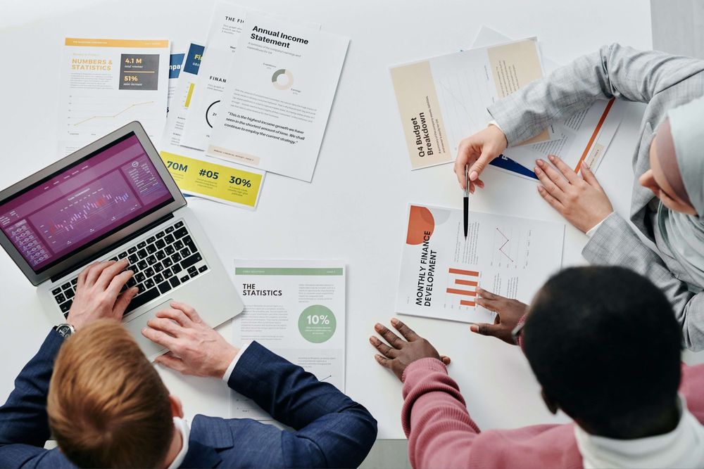 foto superior de mesa com três pessoas analisando relatórios em um computador e várias folhas com gráficos 