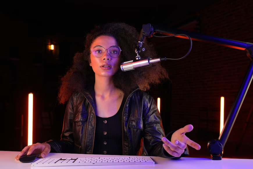 Mulher negra vestindo roupas pretas falando em um microfone prata em frente a mesa com iluminação alaranjada no fundo.