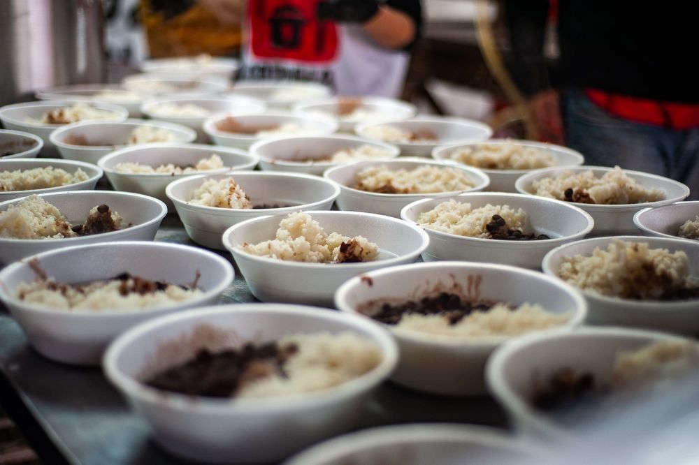 Fotos de diversas marmitas, espalhada em uma mesa de alumínio com uma pessoa ao fundo, desfocada, montando as marmitas.