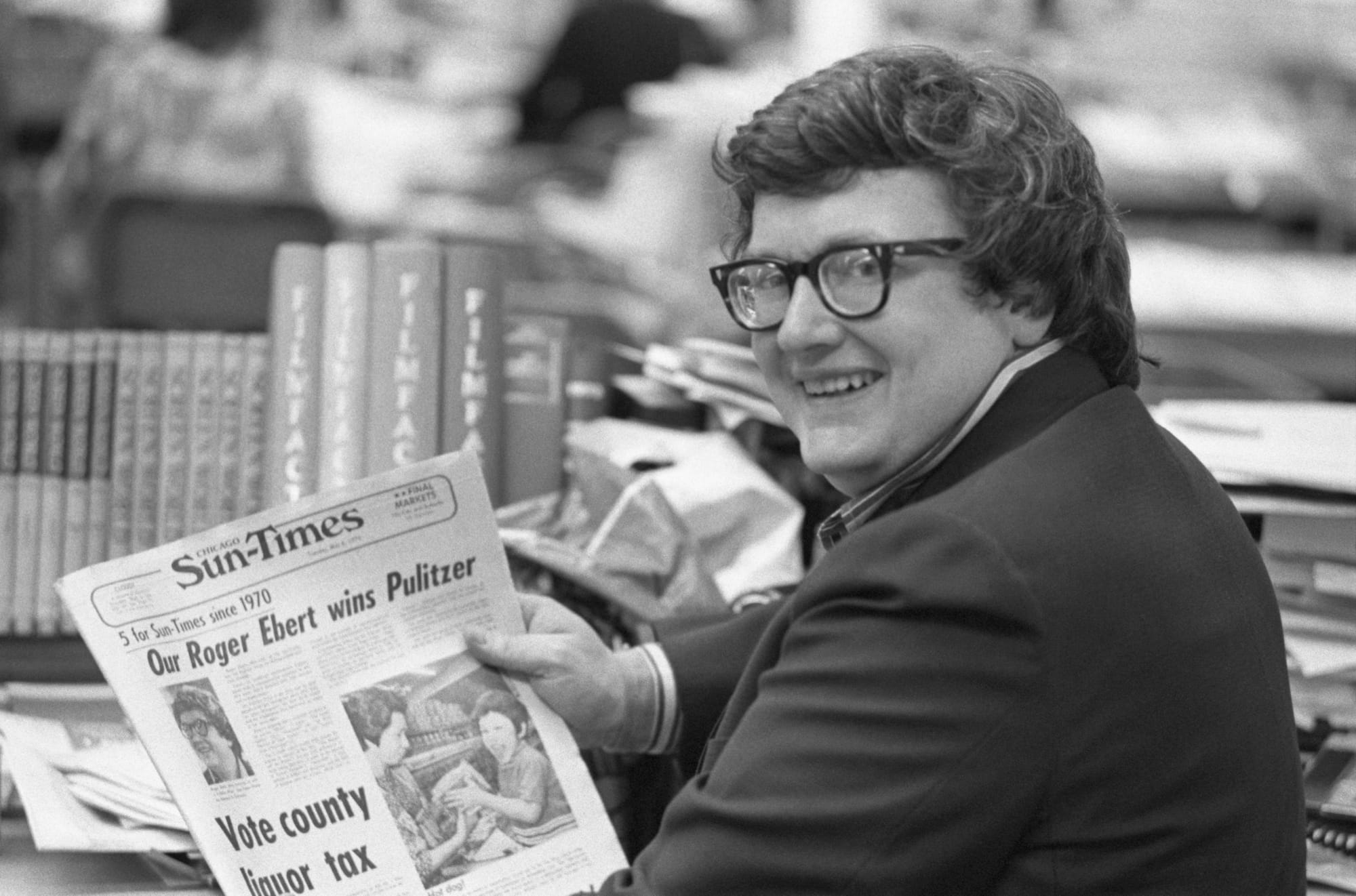 Roger Ebert looks over his shoulder in a black and white photo from 1975 as he sits in a newsroom holding a paper announcing his Pulitzer win.