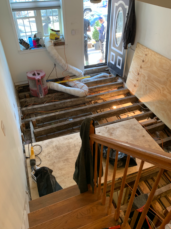 Plywood boards lie across exposed joists between a front door area and a staircase landing where the photographer stands. Below, conduit and basement walls are visible.
