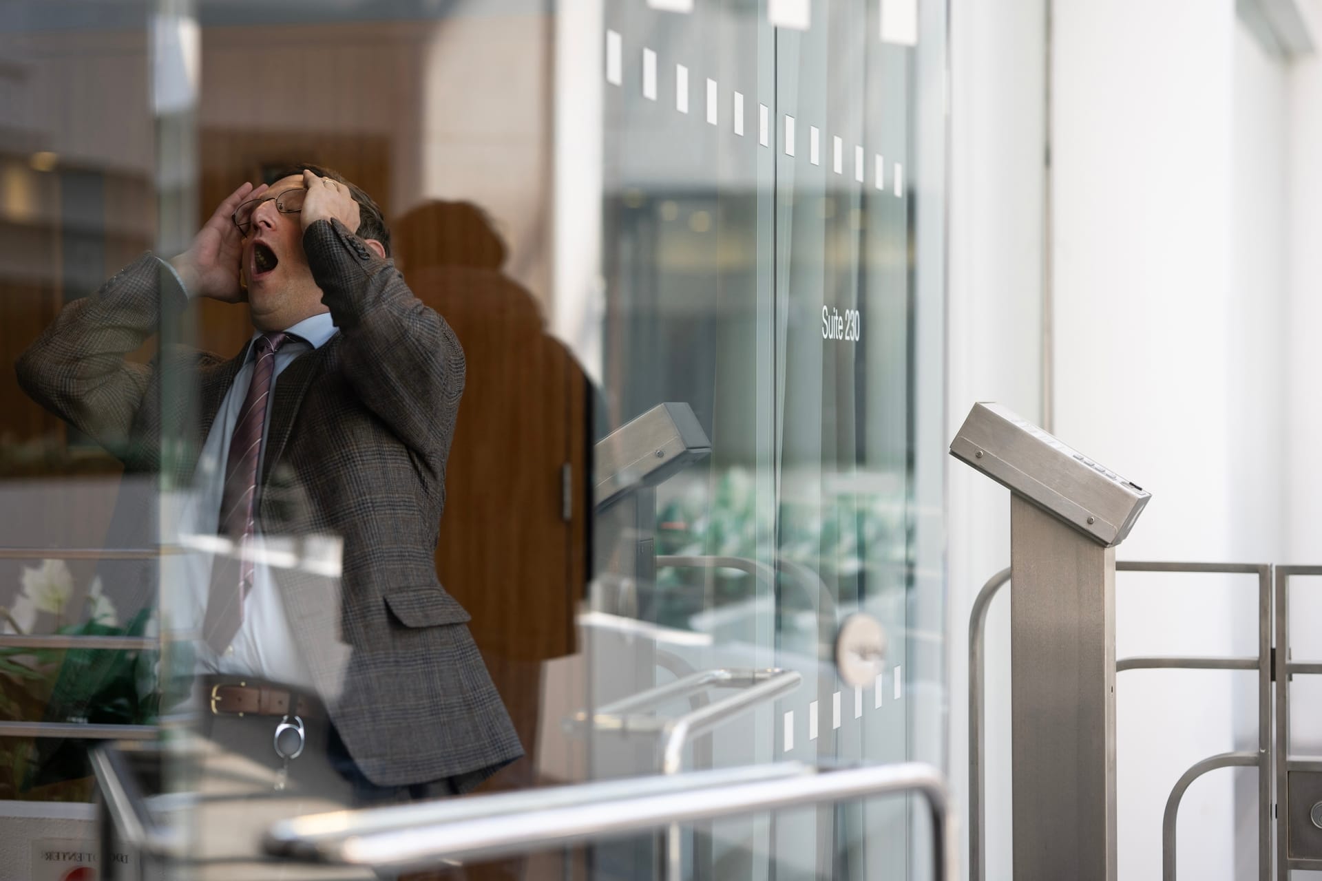 Tim Robinson in an office with glass walls holds his head in his hands while he exclaims in apparent surprise.