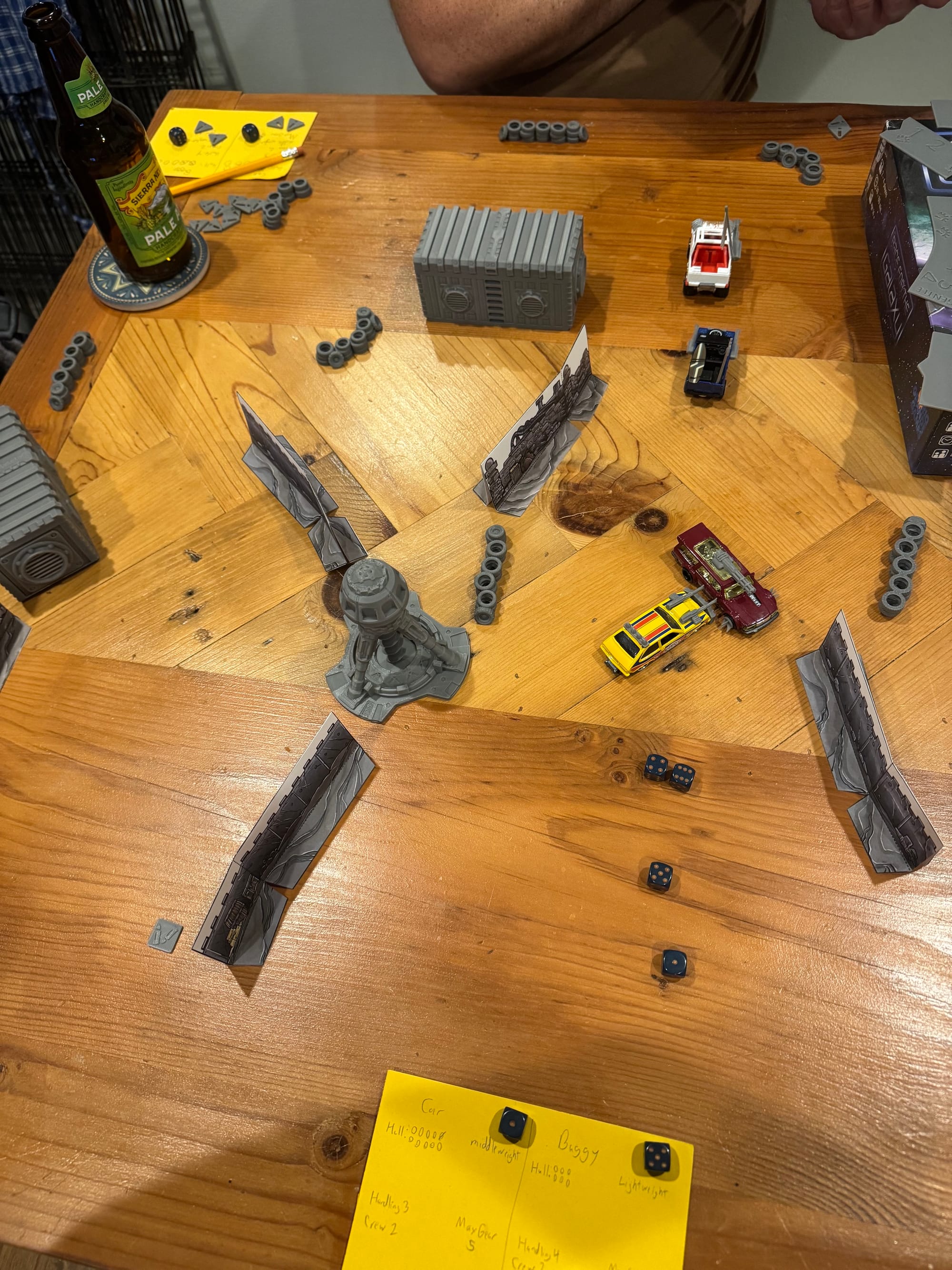 A wood table with 3D-printed terrain littered with dice and hand-written unit information cards, while tiny militarized Hot Wheels-style cars are positioned against one another.
