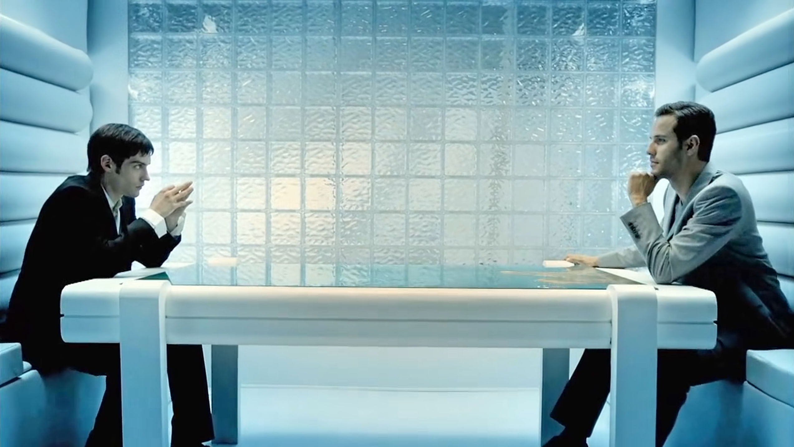 Two models, one representing evil in a gray sportcoat with styled hair and one representing good in a sober dark suit, sit across from one another at a game table.