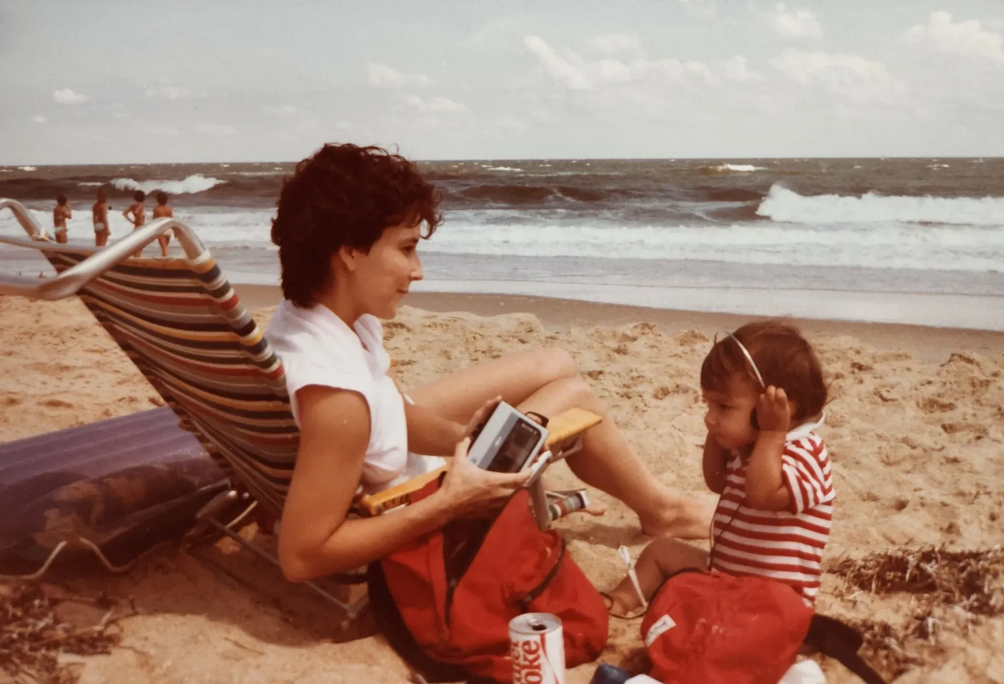 An old photo depicting a woman and child siting on a sandy beach as waves roll in. The woman smiles holding a casette player as the child hunches in concentration with a pair of headphones. The colors are warm, shifted toward red.