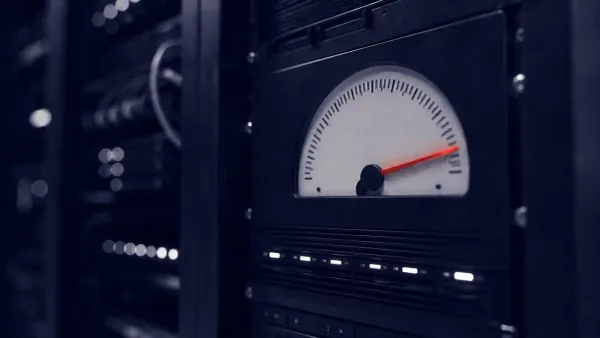 Duotone photo of a dim server room, close on a rack metering panel whose large analog usage gauge has its needle pinned hard to the far end of the dial; only the needle is signal-red.