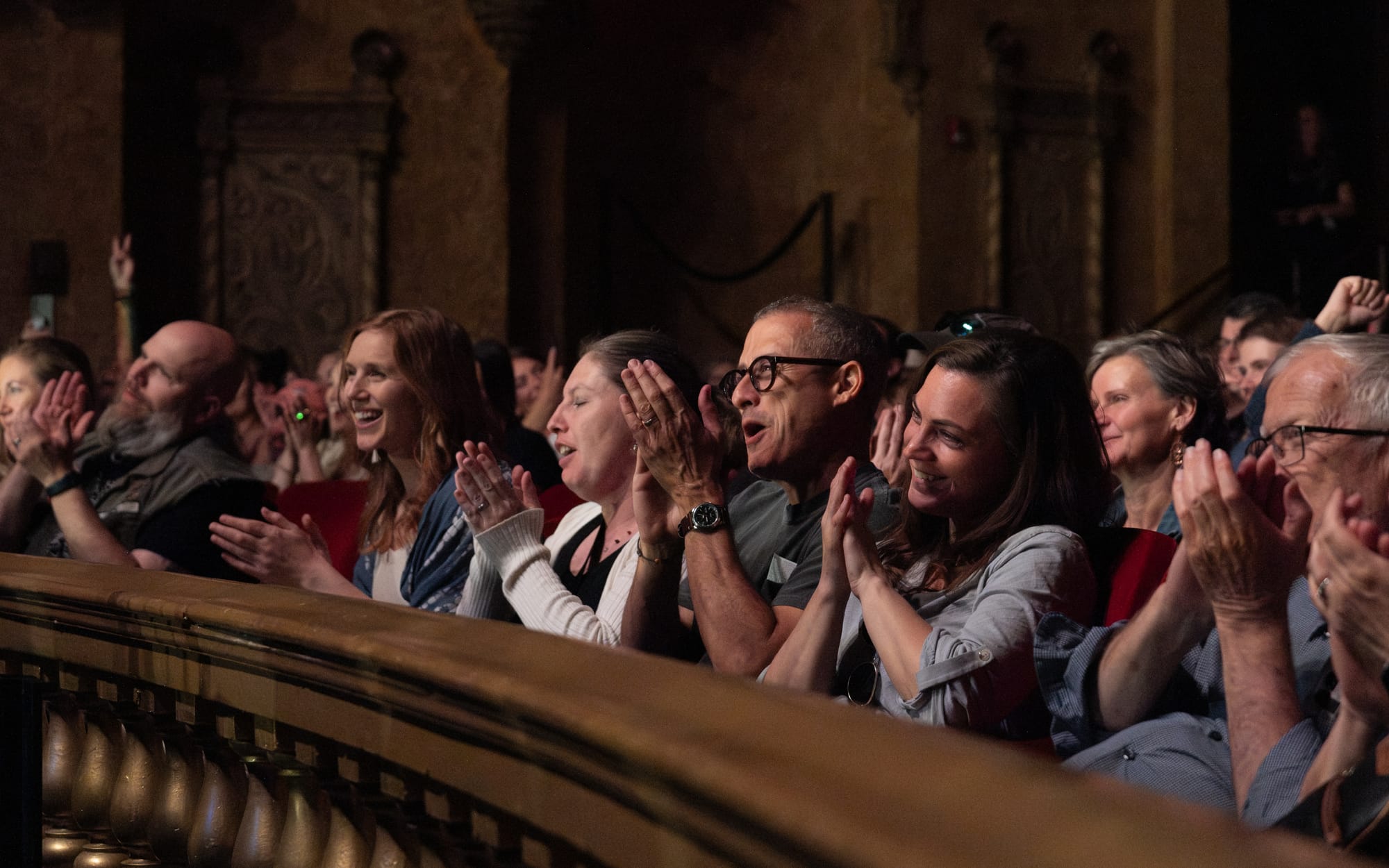 A smiling and clapping audience against a gold barrier.