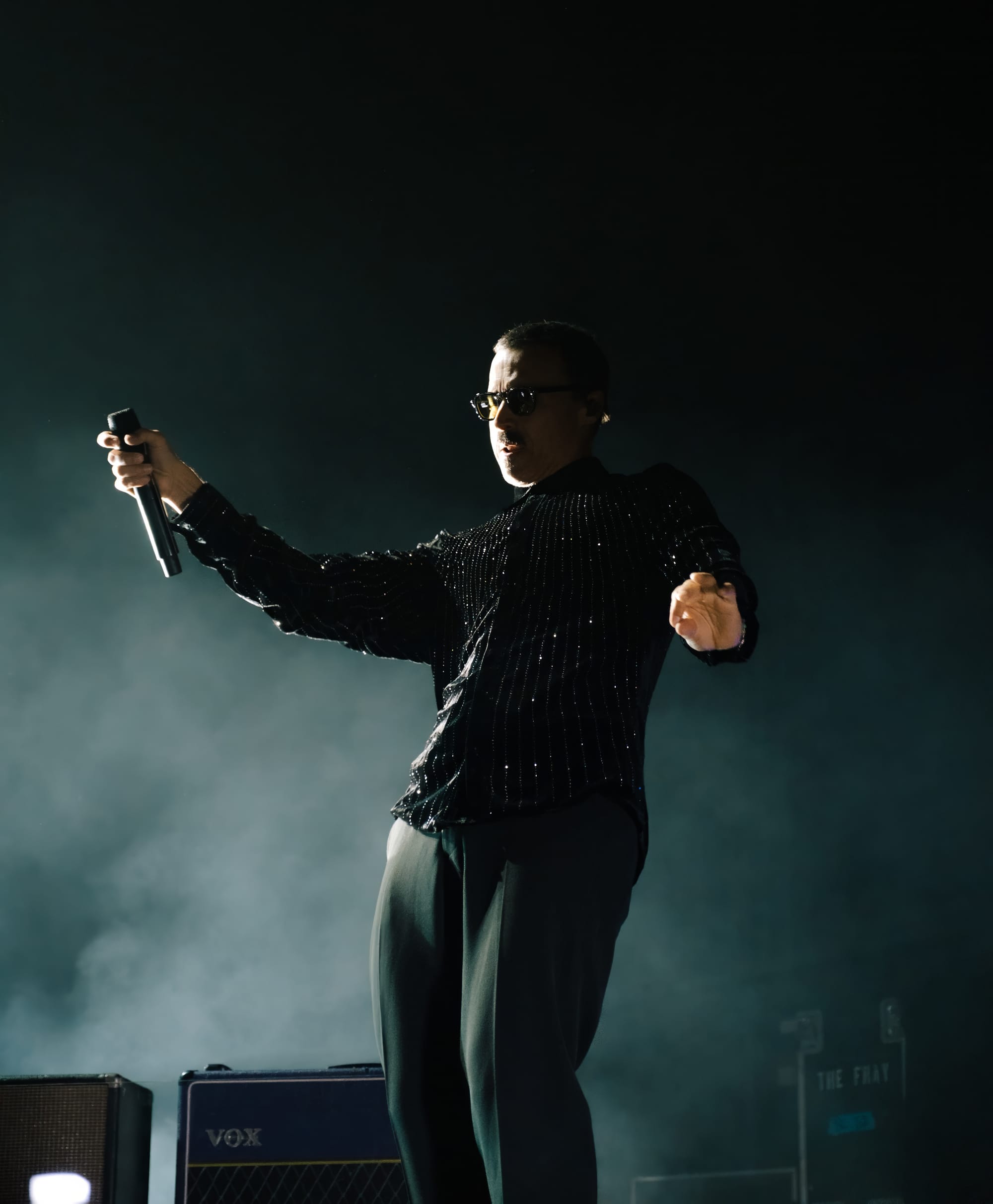King dancing out onto stage as The Fray begins performing. He is wearing a black sequin shirt and slacks, being lit from the side.