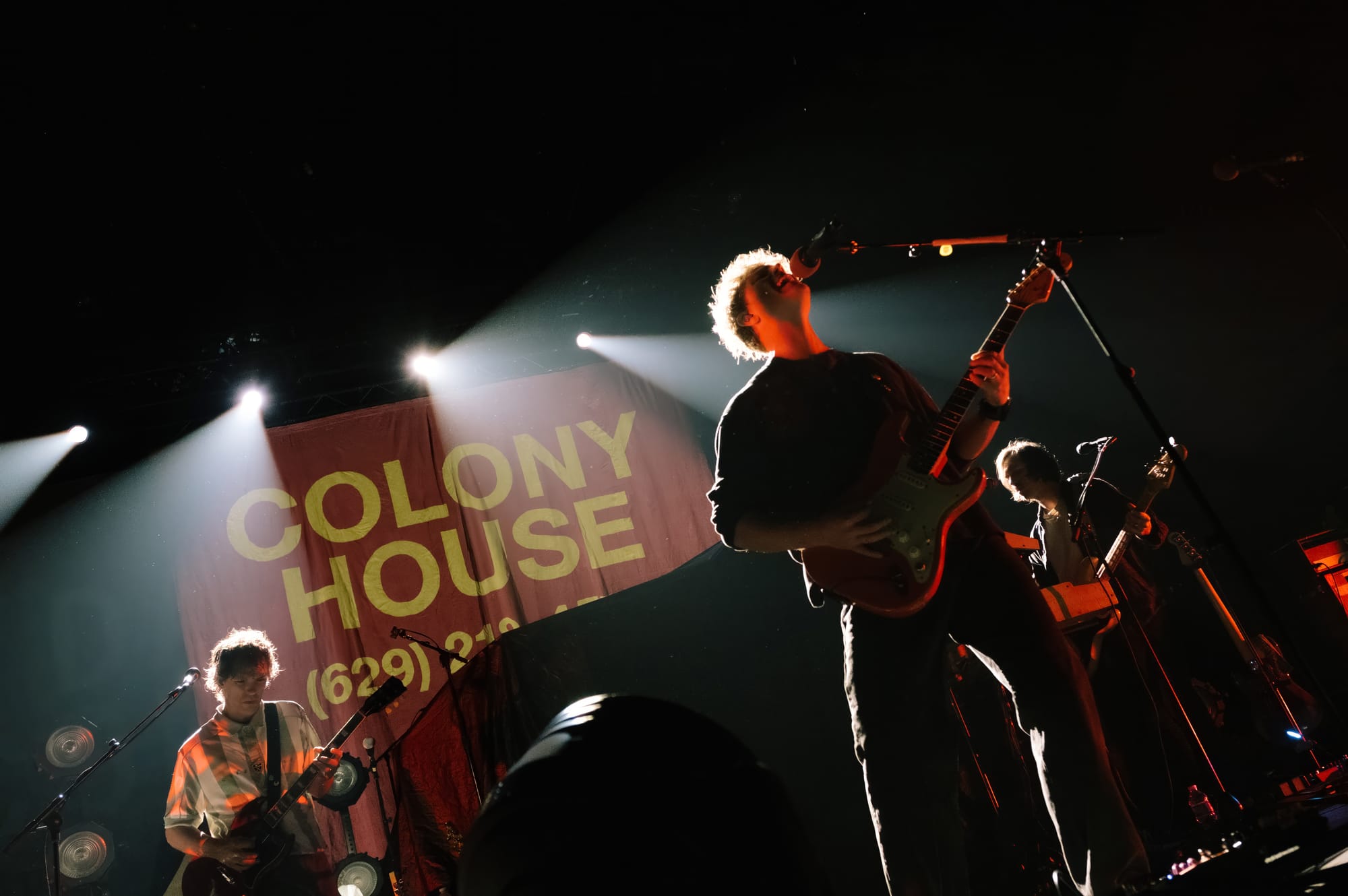 Colony House on a relatively dark stage against a red backdrop reading "Colony House" and a phone number in mustard yellow text. They're backlit by strong hazy stage lighting.