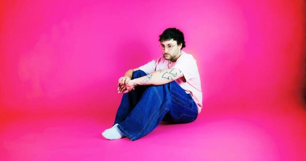 Portrait of artist Reece Young, sitting against a pink studio backdrop and wearing a T-shirt and jeans