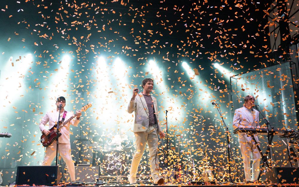 OK Go surrounded by a snowstorm of orange confetti, backlist by pale teal lighting.