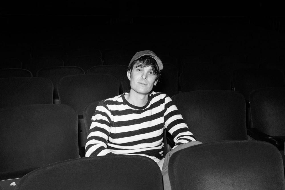 Wes Parker wearing a black and white striped shirt and a baseball cap, looking calmly towards the camera. He is seated in theatre-style seating.