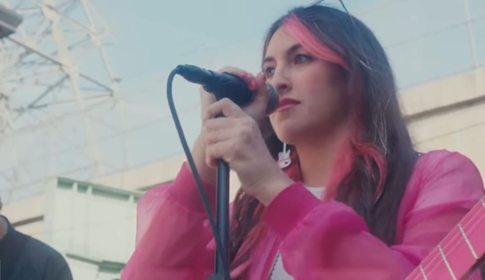 Artist Candice Lee wearing a pink jacket and standing against a microphone, screenshot from a YouTube video