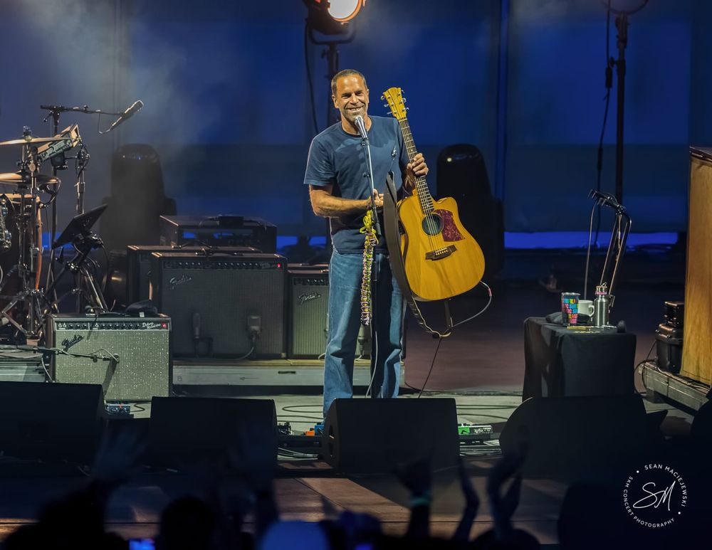Jack Johnson holding up a guitar and standing against a microphone on a stage