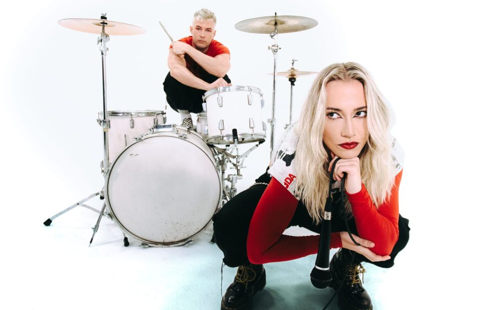 Pop-rock duo senses photographed in an all-white studio, with vocalist Madison Taylor crouching with a microphone in the foreground and drummer Nick Sampon in the background with a drumset