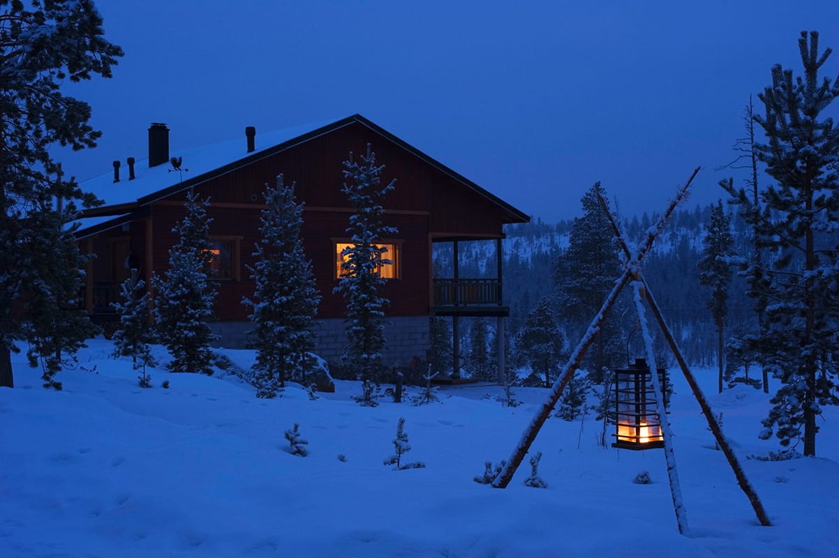 Snow-covered cabin at dusk. A window is lit up from inside, and in the foreground are three logs tipped together in a pyramid with a hanging lamp of fire between them.